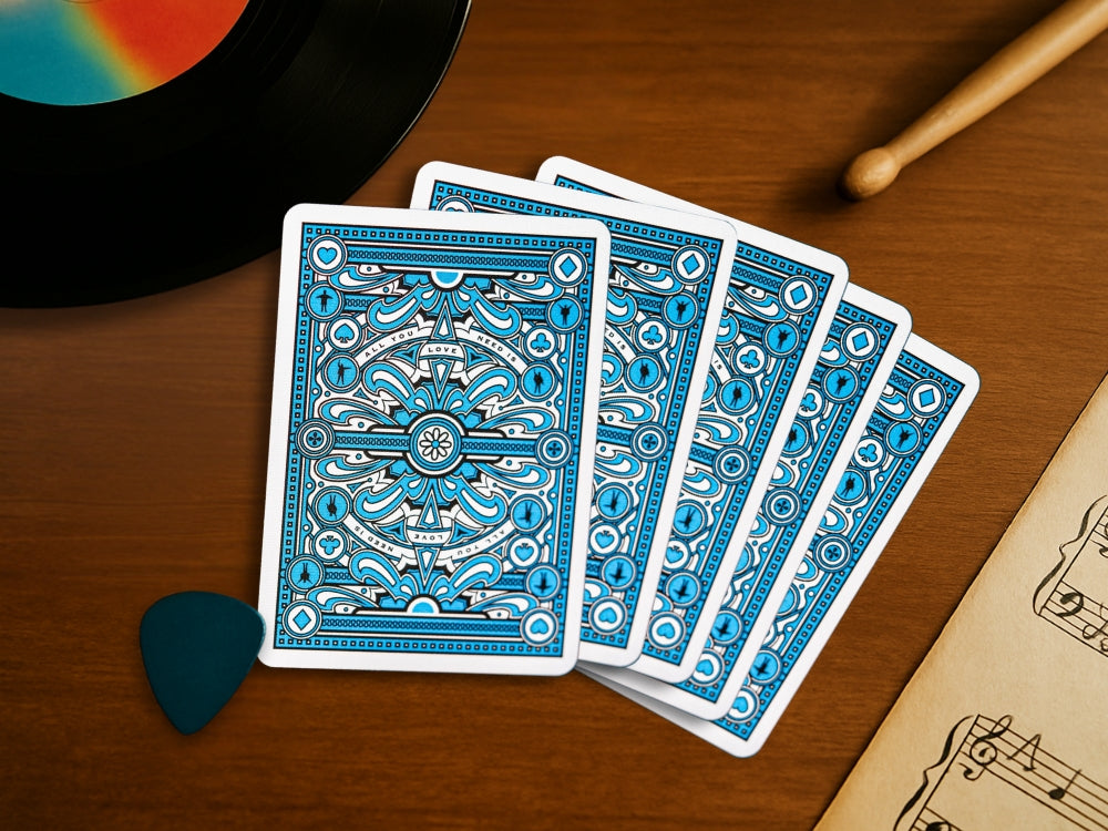 A fan of five blue-backed theory11 playing cards lies on a wooden table, surrounded by musical elements including a vinyl record, guitar pick, drumstick, and sheet music - visually tying into the theme of songs about playing cards.