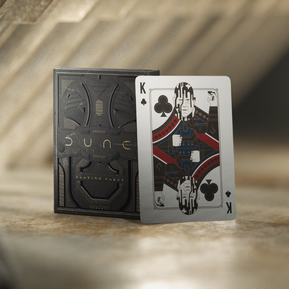 King of clubs playing card, leaning against the dune playing cards deck box. all on a sandy coloured background.