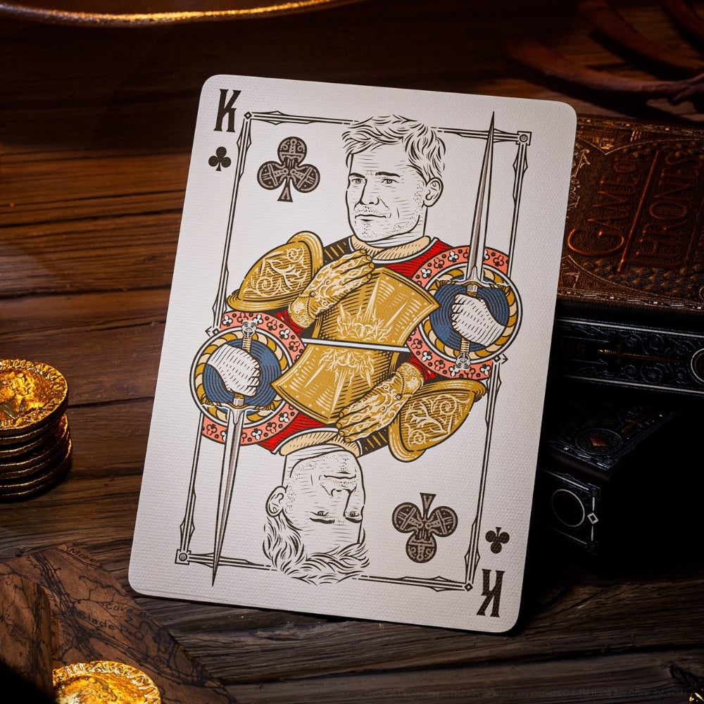 King of clubs taken from a deck of Game of thrones playing cards. The card is sitting on a wooden table, leaning against two deck boxes, with gold coins stacking near by