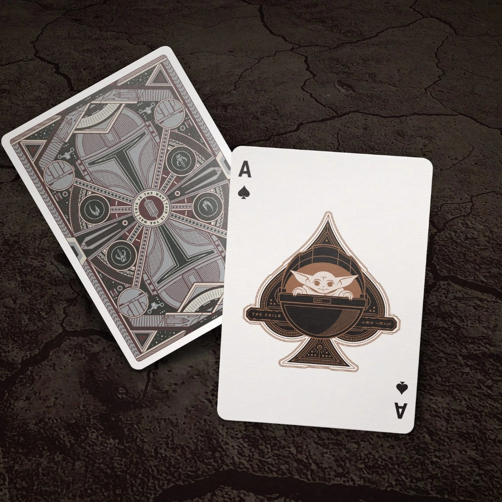 an ace of spades card featuring Grogu from the mandalorian playing cards deck, laying over a facedown card, both on top of a cracked and dry ground.