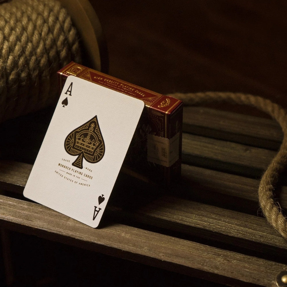 Ace of spades from a deck of Red Monarch playing cards.  The card is leaning against a deck box on a slatted wooden surface.