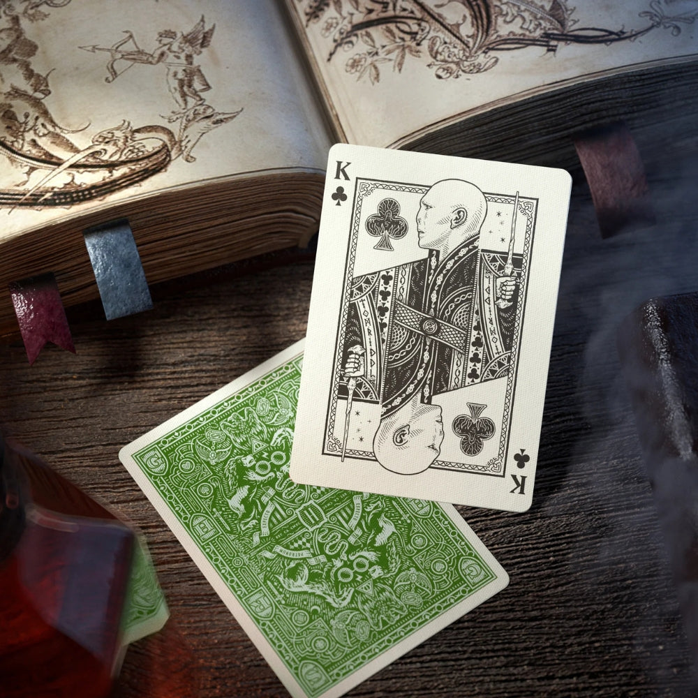 King of clubs laid over a face down card from the Slytherin pack of playing cards and leaning against an old book.
