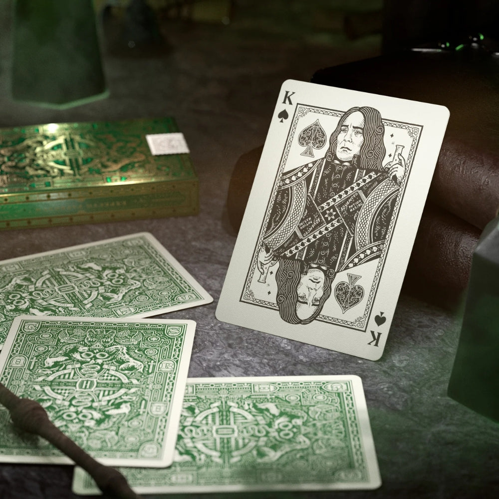 king of spades leaning against two leather bound books, with three Slytherin harry potter playing cards laying face down.  There is a Slytherin playing cards deck box in the background.